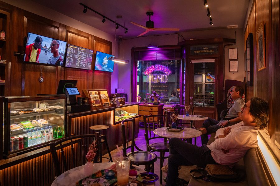 ลูกค้านั่งชิลที่โต๊ะคาเฟ่ ใต้ทีวีเมนูและไฟนีออนสีม่วง